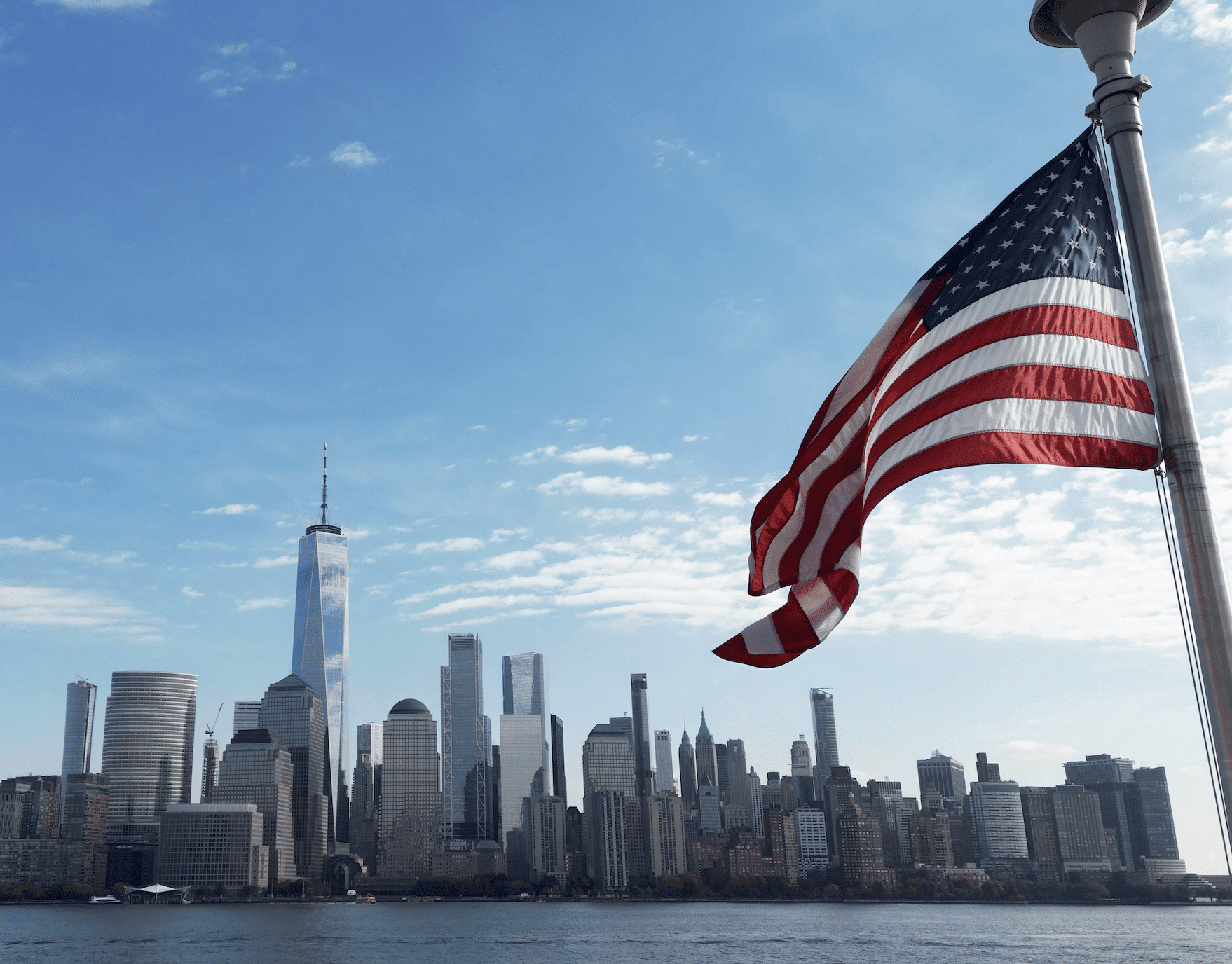 An American flag waves in the foreground with a city skyline and body of water in the background.